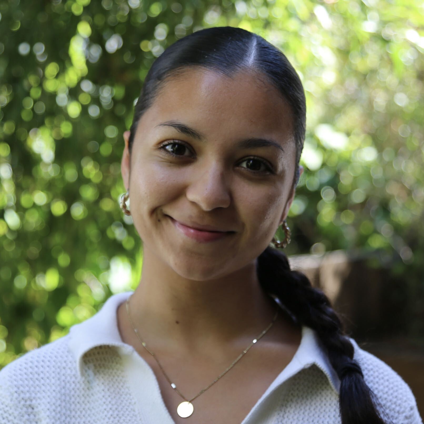 A woman with dark hair in a braid, wearing a white top and gold jewelry, stands outdoors in front of green foliage, smiling slightly.