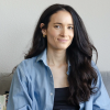A woman with long dark hair wearing a blue shirt and gold hoop earrings sits on a gray couch, smiling slightly at the camera.